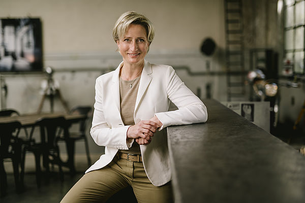 Dr. Nicole Hoffmeister-Kraut ist Schirmherrin des AI Communication Award Dr. Nicole Hoffmeister-Kraut lächelnd neben einer Bar auf die sie den Arm lehnt